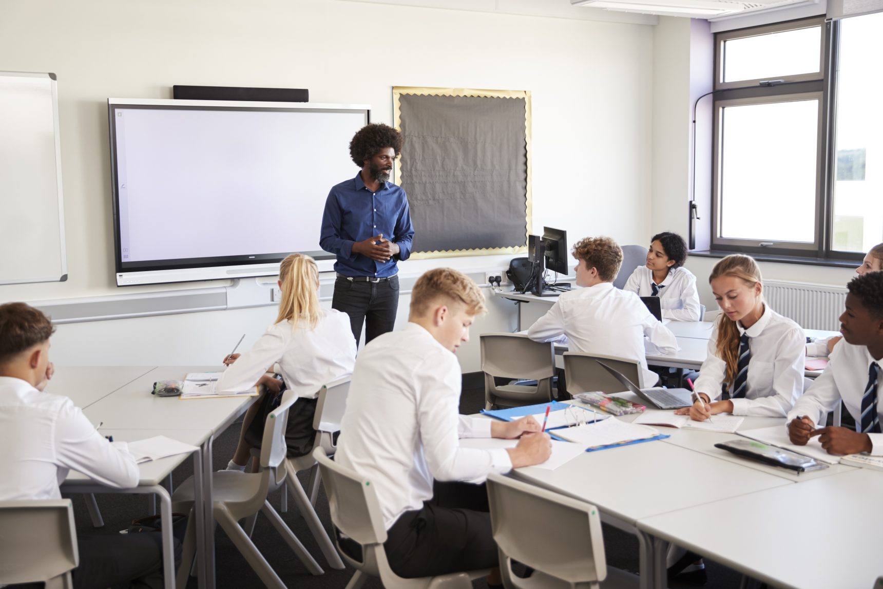 A male secondary school teacher standing next to an interactive whiteboard and teaching a lesson to his pupils.