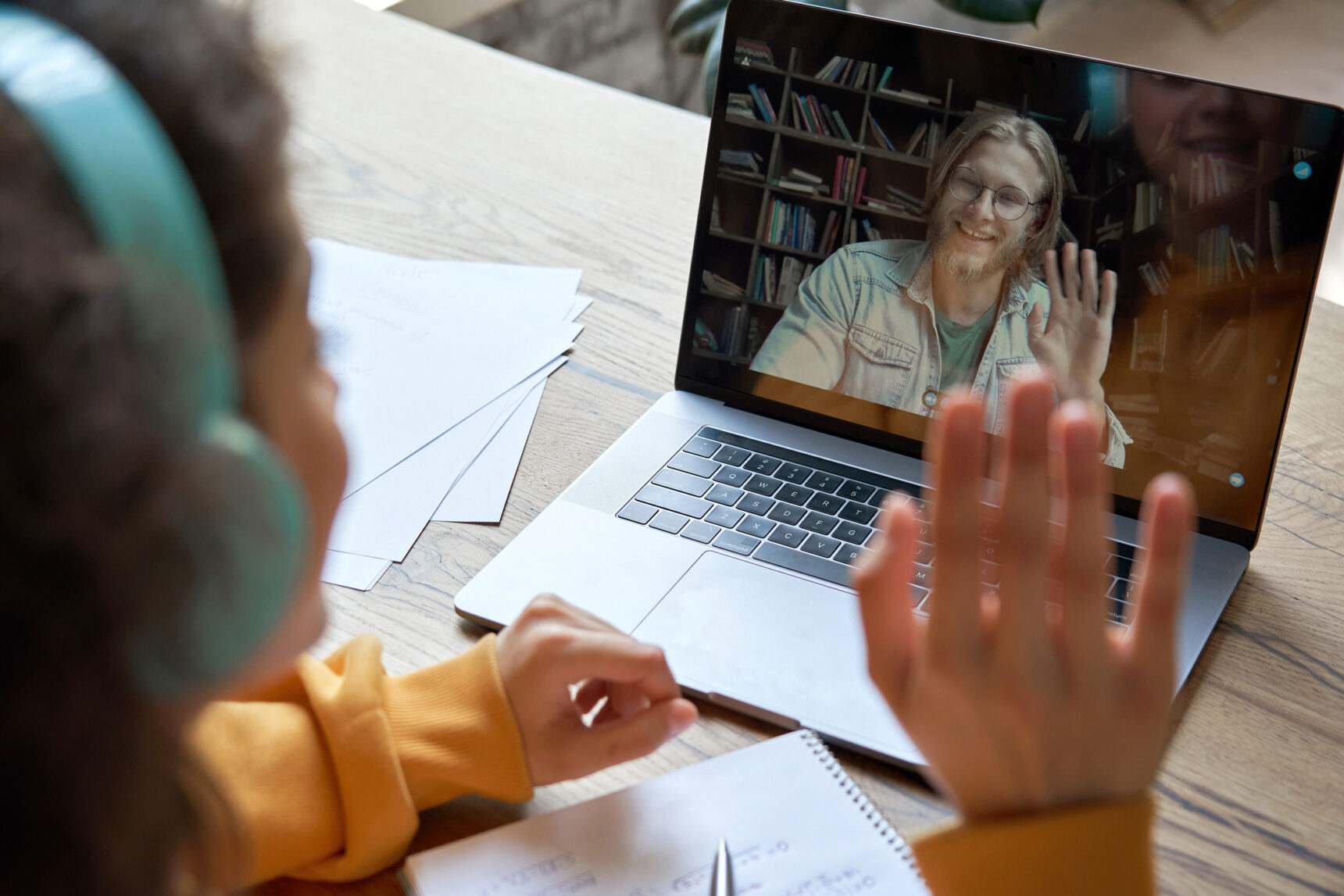 A young student is waving at her online tutor during an online tutoring session on her laptop.