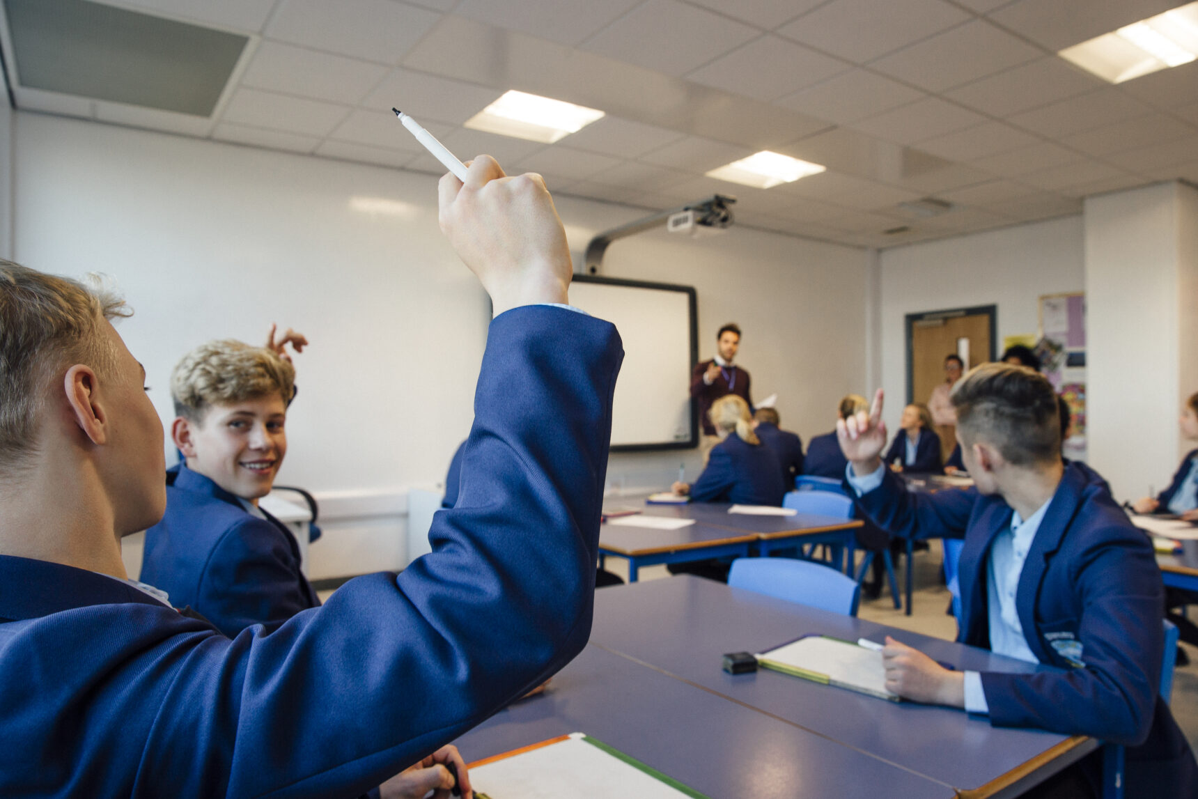 A classroom of students with mini whiteboards on the desks are raising their hands to answer the teacher's question.