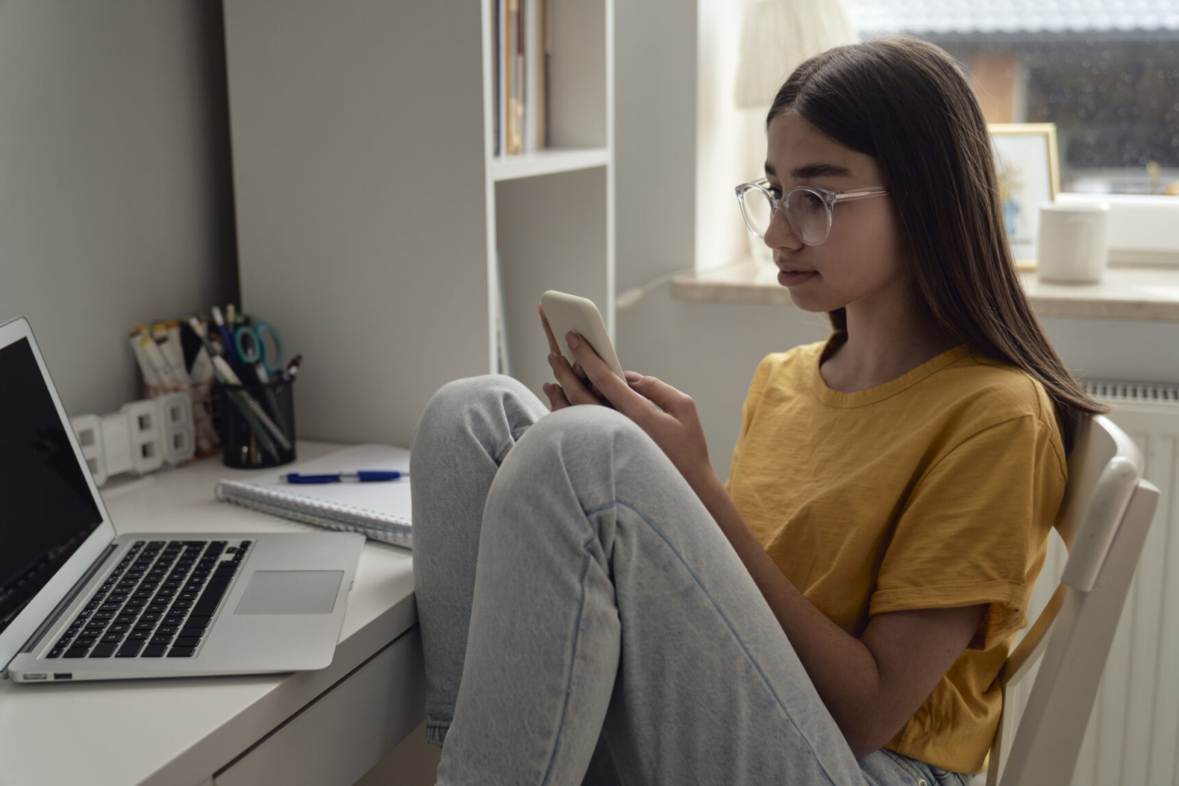 A teenage girl is distracted by her smartphone while studying at a desk.