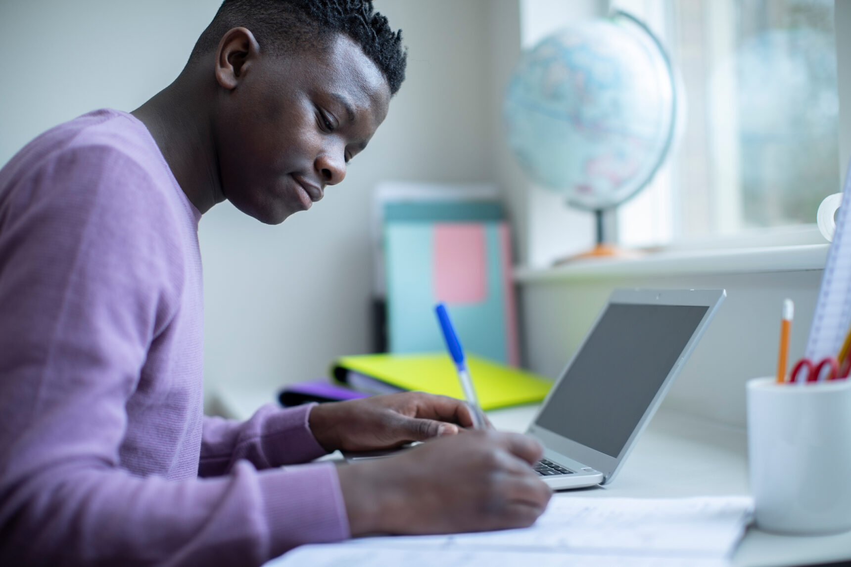 A teenage boy is sitting at a desk with a laptop, taking notes as he prepares for the GCSE English Language exam.